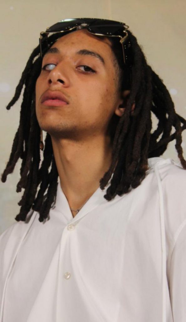 Modelo da Fenty, homem negro, magro, sem barba, usando uma camisa branca, com dreads no cabelo, e visão monocular.