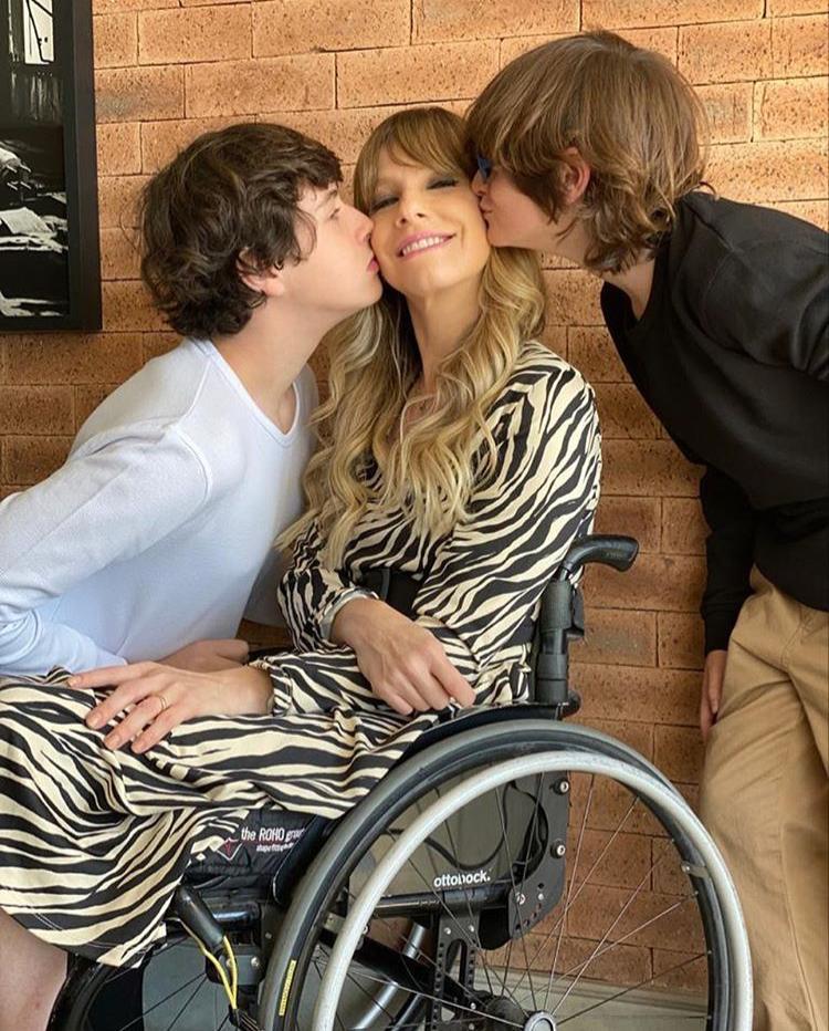Foto de uma mulher sentada na cadeira de rodas, usando um vestido sendo beijada nas bochechas por seus dois filhos, um a sua direita, e um a sua esquerda. Ela sorri,