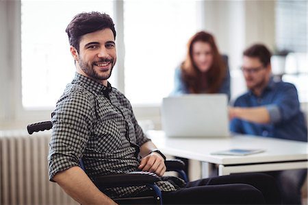 Homem sorrindo na cadeira de rodas, ao fundo, uma mulher e um homem usado um notebook. Foco da imagem na diversidade