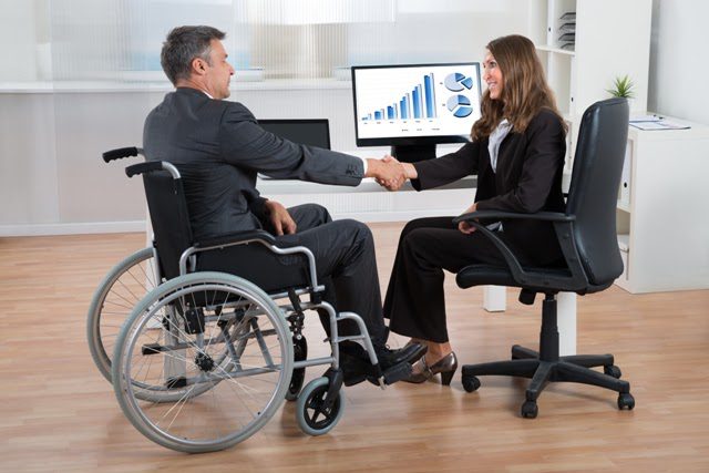 à esquerdad da imagem está um homem em uma cadeira de rodas, usando terno, apertando a mão de uma mulher com cabelo castanho sentada em uma cadeira de escritorio. Ela sorri para o homem, há um computador com gráficos que expõe dados da liderança da empresa