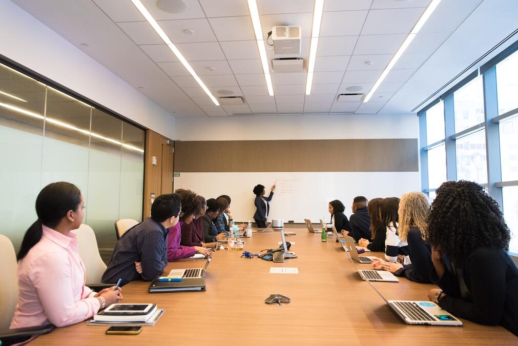 Funcionários sentados em mesa de reunião com seus laptops e agendas e líder expondo informações em quadro diante deles