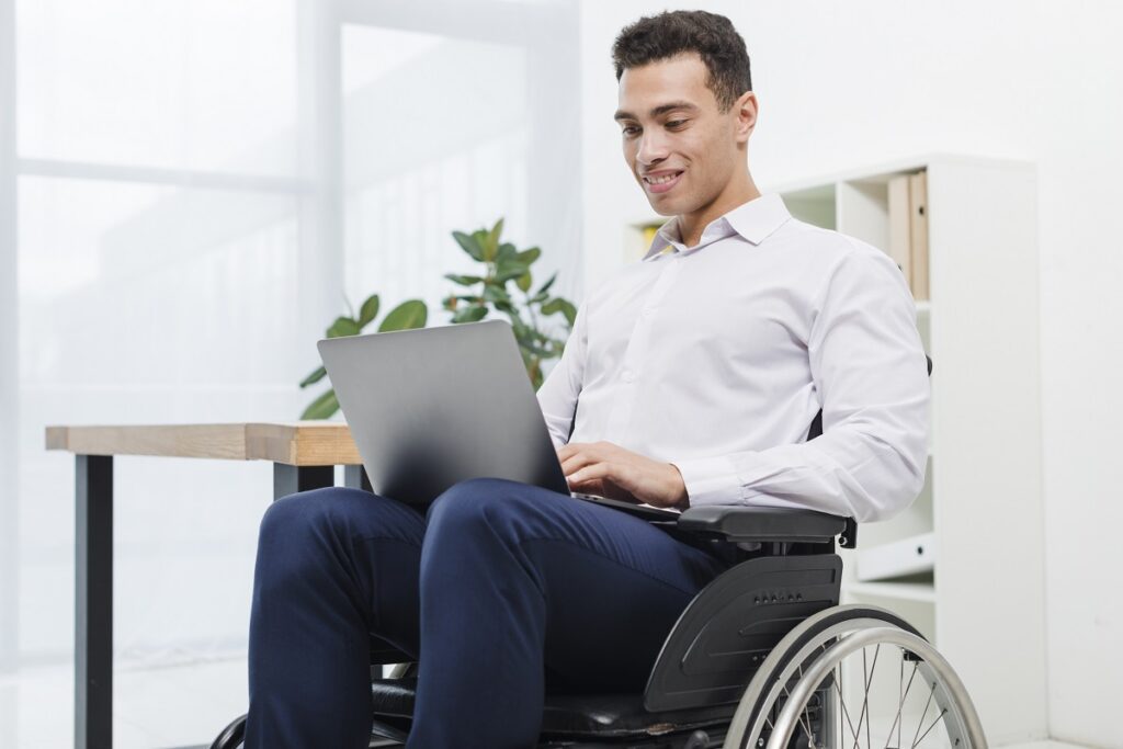 Homem jovem sorrindo em cadeira de rodas com notebook no colo e ao fundo mesa com jarro de plantas.