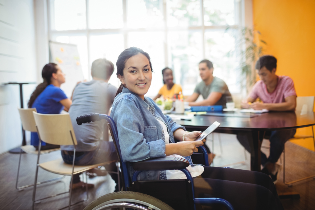 Mulher em cadeira de rodas sentada sorri para a câmera com tablet na mão. Atrás dela, colegas de trabalho sentados um diante do outro em uma mesa.