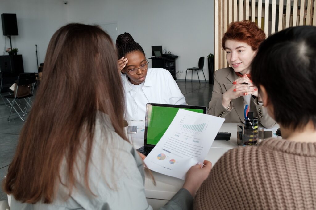 Reunião de mulheres em trabalho, de diversas etnias, enquanto analisam dados em relatório, com laptop sobre a mesa