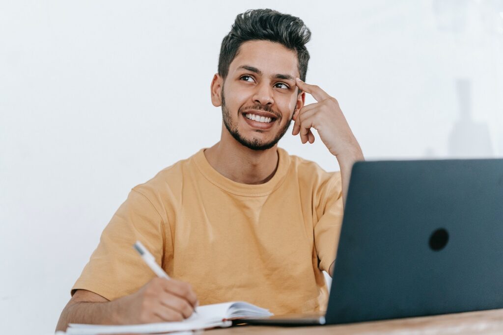 Homem sorridente e pensativo, sentado em cadeira de trabalho diante de seu laptop, enquanto escreve em agenda