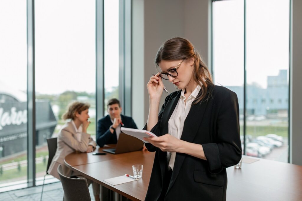 Pessoas em local de trabalho vestindo blazers, uma mulher em pé de óculos analisando dados e casal sentado mais a fundo com notebook à sua frente