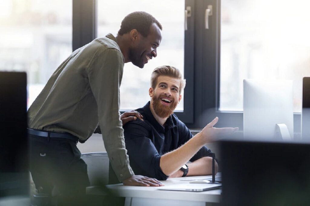 Dois homens sorridentes, um em pé e outro sentado, enquanto analisam algo no computador.