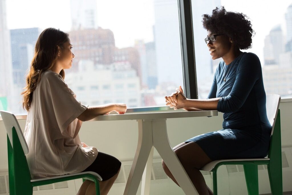 Mulheres sentadas em mesa uma de frente à outra em ambiente de trabalho.