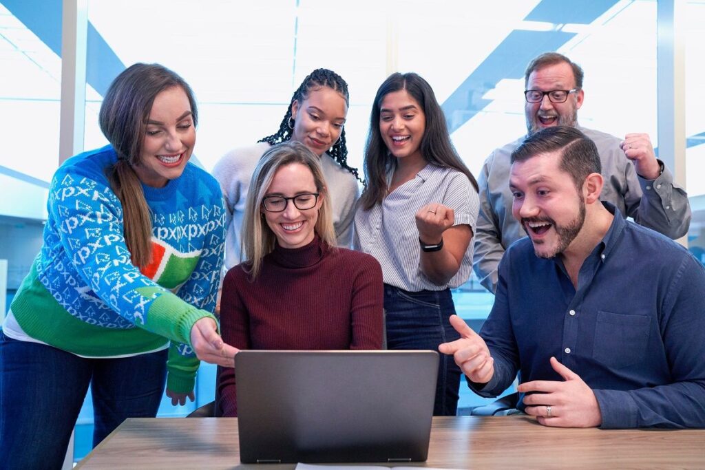 Homens e mulheres em ambiente de trabalho ao redor de computador enquanto sorriem.