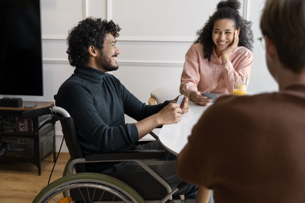 Pessoas conversam em mesa no ambiente de trabalho, uma mulher de costas e uma de frente sorri enquanto conversa com homem também sorridente em cadeira de rodas.