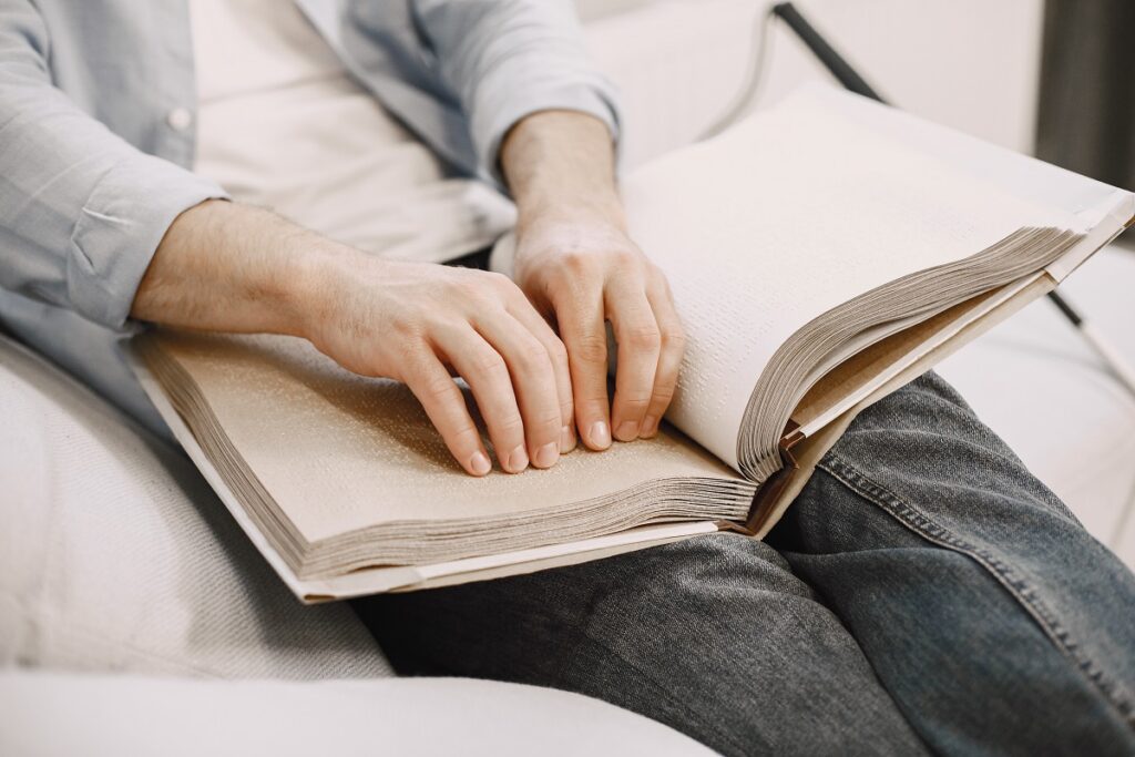 Pessoa sentada lê livro em braille, no detalhe da imagem ele está sentado com o livro sobre as pernas