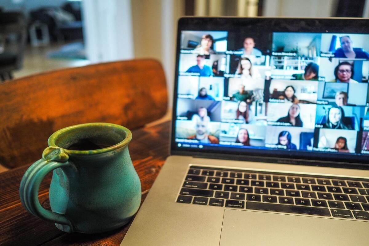 Imagem de uma tela de computador onde acontece uma reuniao online com várias pessoas logadas, na mesa tem uma caneca azul