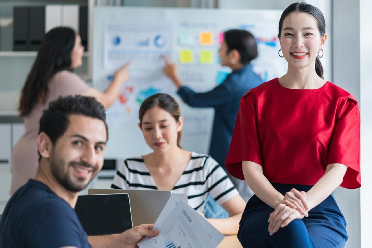 Uma sala com várias pessoas trabalhando em equipe. Duas delas conversam em um quadro com esquemas e post-its, outra digita ao computador e e duas olham para frente sorrindo