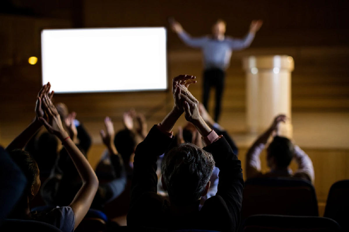 Pessoas na plateia aplaudindo um palestrante que está no palco