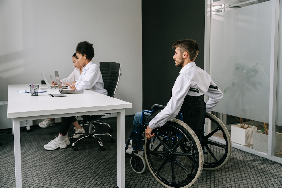 homem em cadeira de rodas, chegando em uma mesa com outras duas pessoas trabalhando