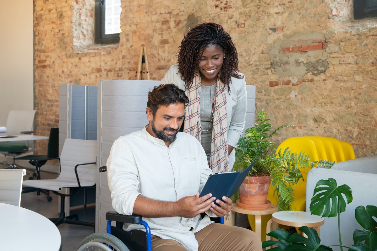Homem cadeirante com tablet na mão com mulher ao seu lado que sorri em ambiente de trabalho análogo à escritório com mesas e computadores ao fundo e ao redor dos mesmos na foto