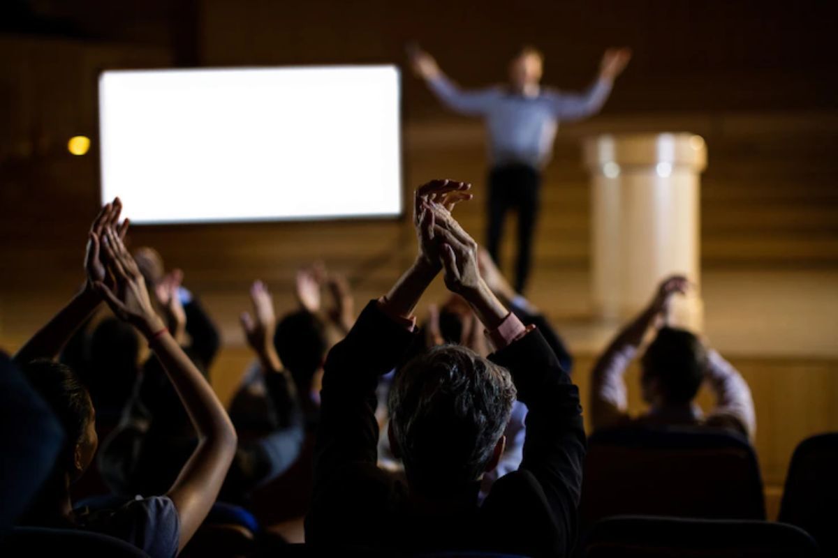 Foto tirada de plateia que aplaude palestrante em desfoco no palco de local análogo à teatro