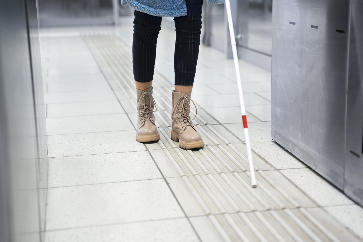 Mulher deficiente visual se desloca com auxílio de seu bastão branco com tira vermelha em ambiente interno de empresa com piso sinalizado