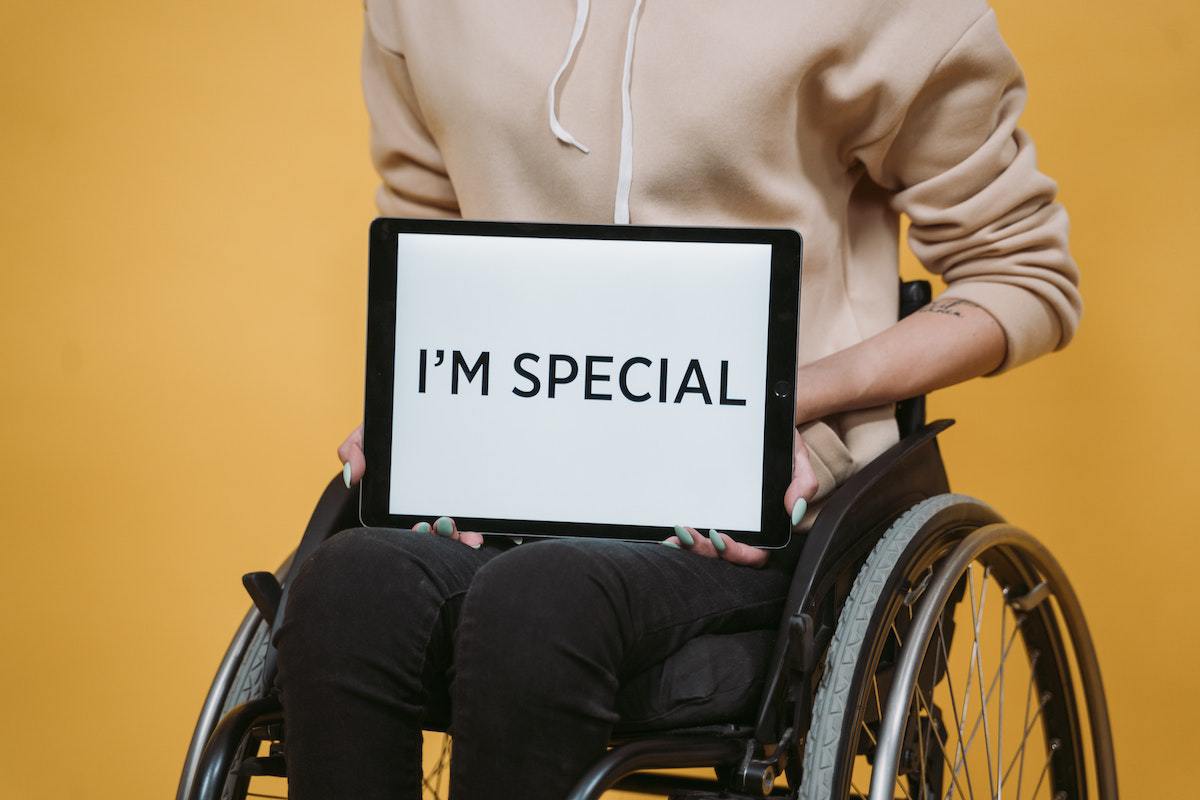 Homem em cadeira de rodas vestido formalmente enquanto segura placa que diz "eu sou especial" escrita em inglês e parede laranja ao fundo