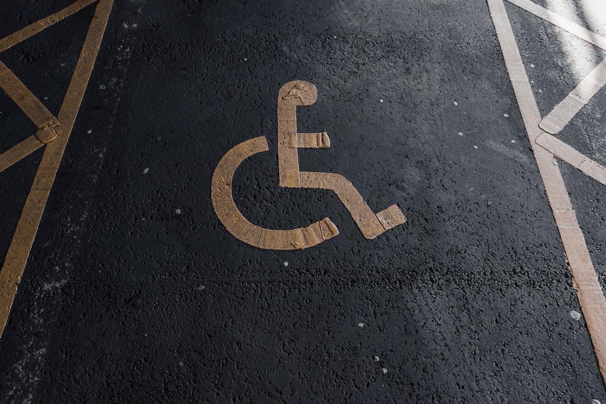 Local análogo à estacionamento com espaço reservado e símbolo de pessoa com deficiência gravado no chão com tinta de cor amarela sobre o piso cinza escuro, quase preto