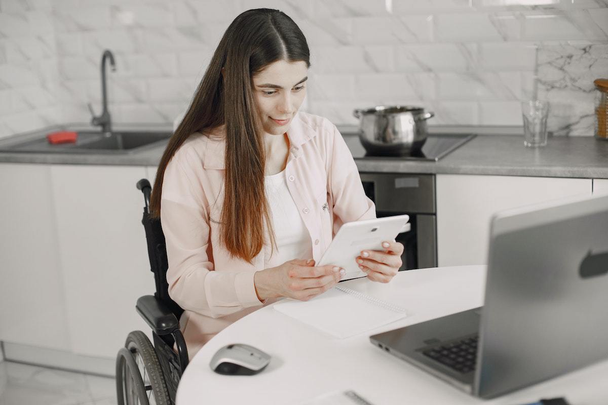 Mulher em cadeira de rodas com tablet na mão e diante de mesa branca redonda com notebook cinza em cima dela e com artigos de cozinha ao fundo