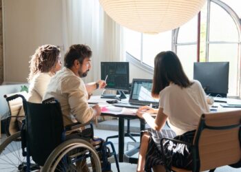 Homem em cadeira de rodas conversando com duas mulheres em local de trabalho diante de seus computadores e sentadas em suas cadeiras