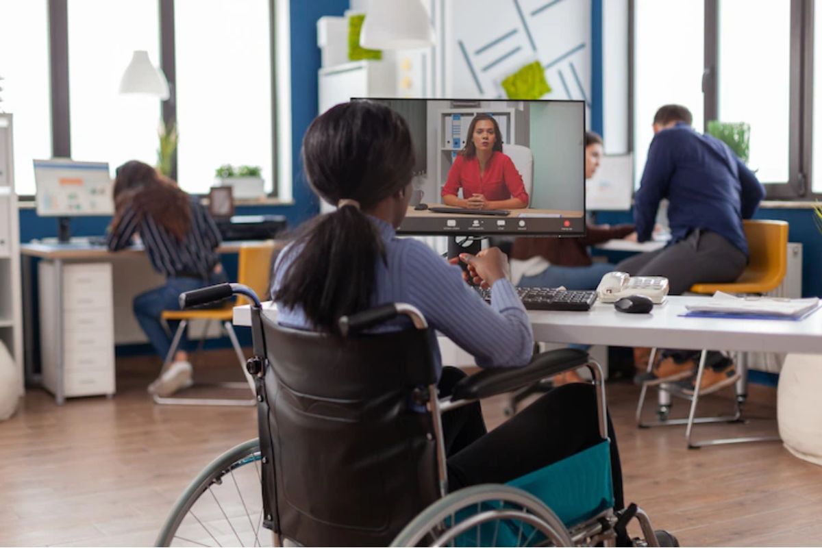 Mulher cadeirante diante de mesa em local de trabalho enquanto assiste algo em seu computador de mesa com outros colegas ao fundo da imagem