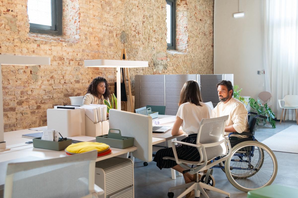 Homem em cadeira de rodas conversando com duas mulheres em escritório com empresa que pratica ações de diversidade e inclusão diante de parede de tijolos dialogando