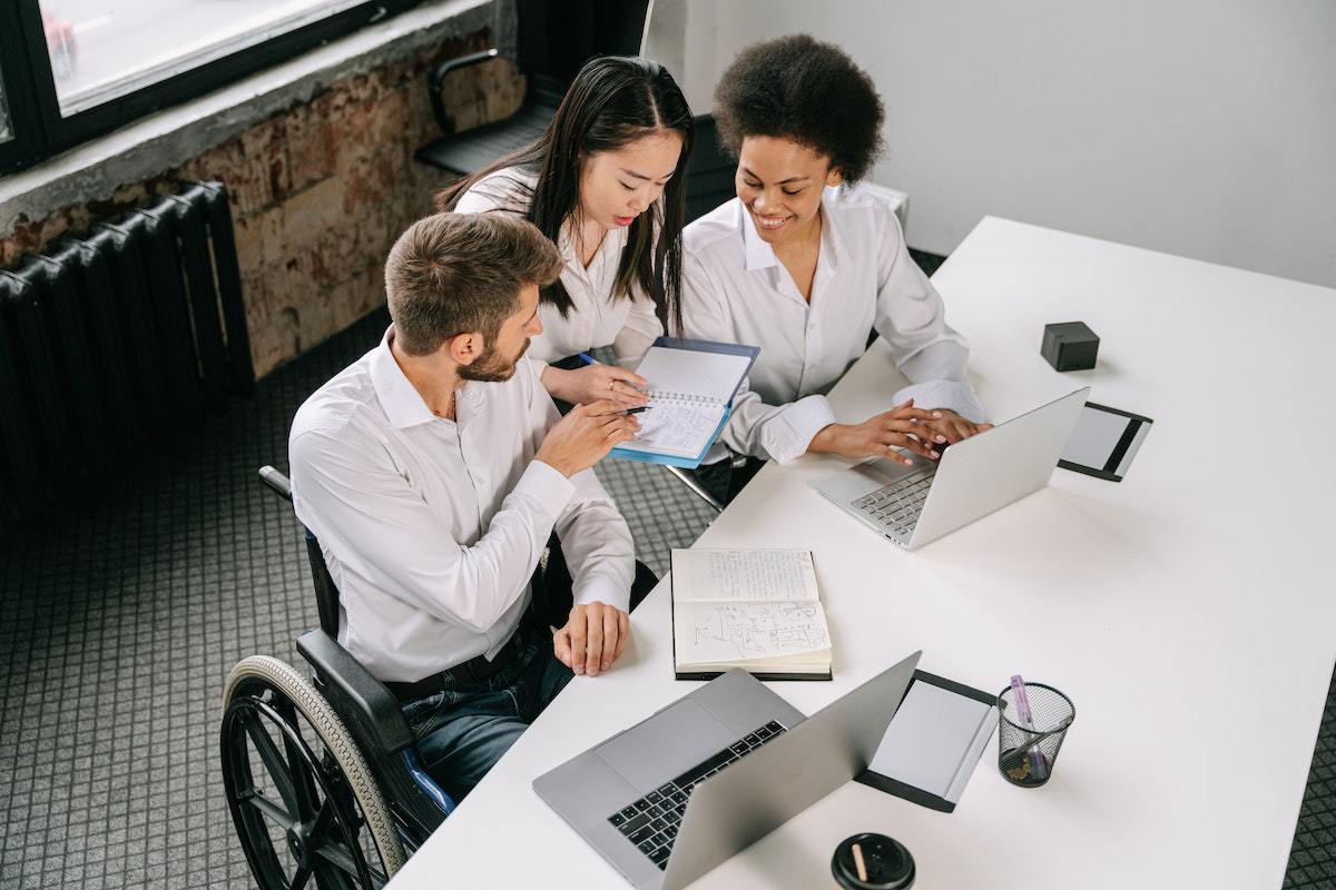 Homem em cadeira de rodas conversando com duas mulheres em escritório com empresa que consegue Aplicar a diversidade