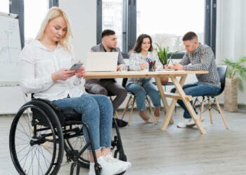 Mulher em cadeira de rodas se preparando para Entrevista de emprego com três pessoas ao fundo em local de empresa inclusiva