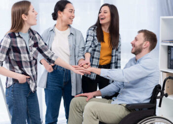 Homem em cadeira de rodas cumprimentando três colegas mulheres de pé em local de trabalho como exemplo de inclusão