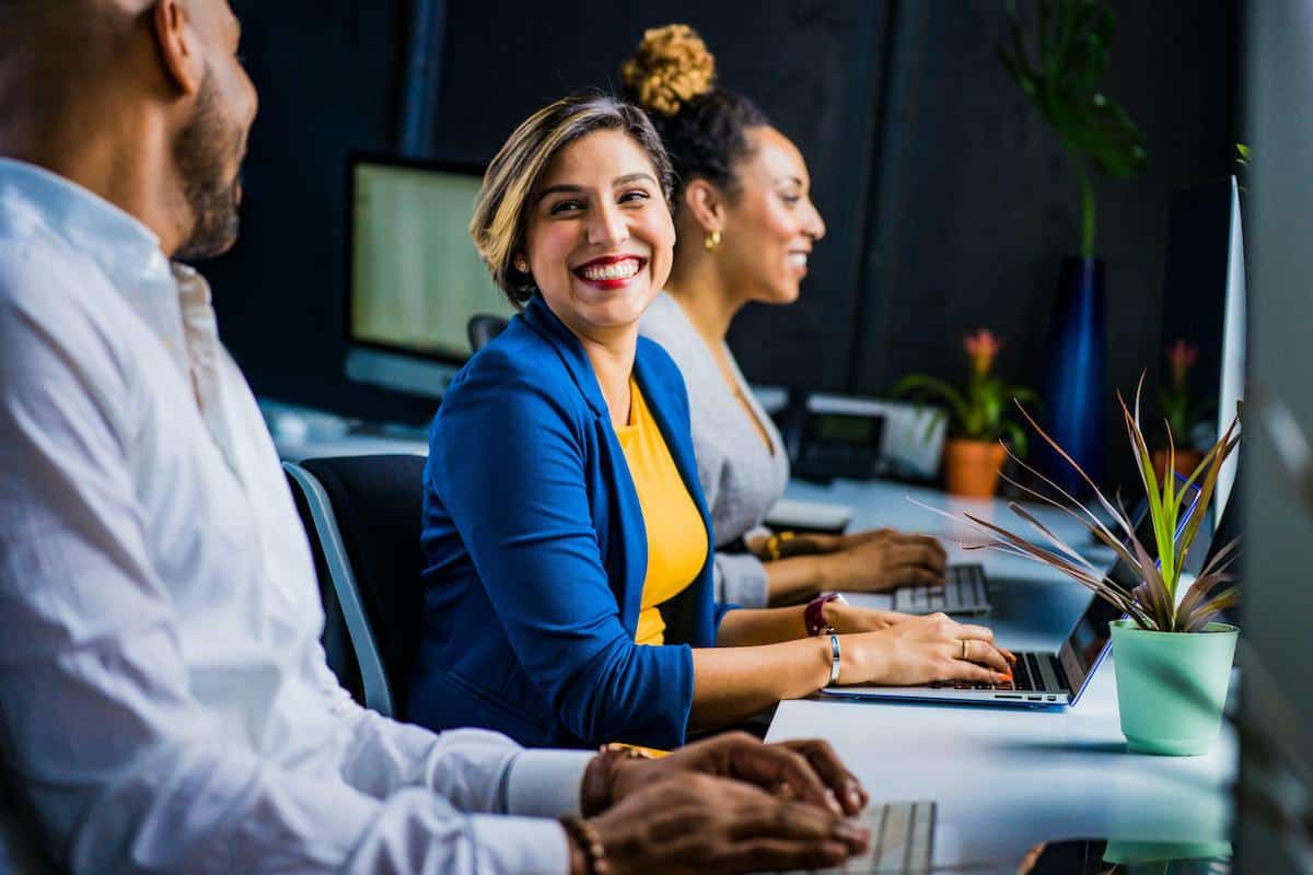 Duas mulheres e um homem sorridentes usando notebooks em local de trabalho enquanto dois se olham como comunicação interna