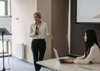 Mulher de pé com microfone realizando Palestra sobre diversidade nas empresas com mulher ao lado sentada usando notebook