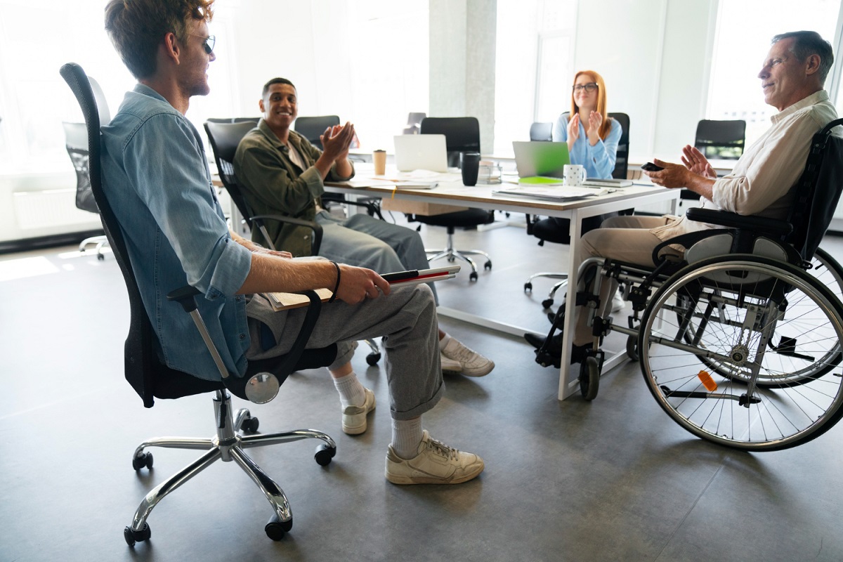 Homem em cadeira de rodas em empresa com Plano de inclusão conversando com dois homens e uma mulher ao redor de mesa em escritório enquanto duas pessoas aplaudem a conversa
