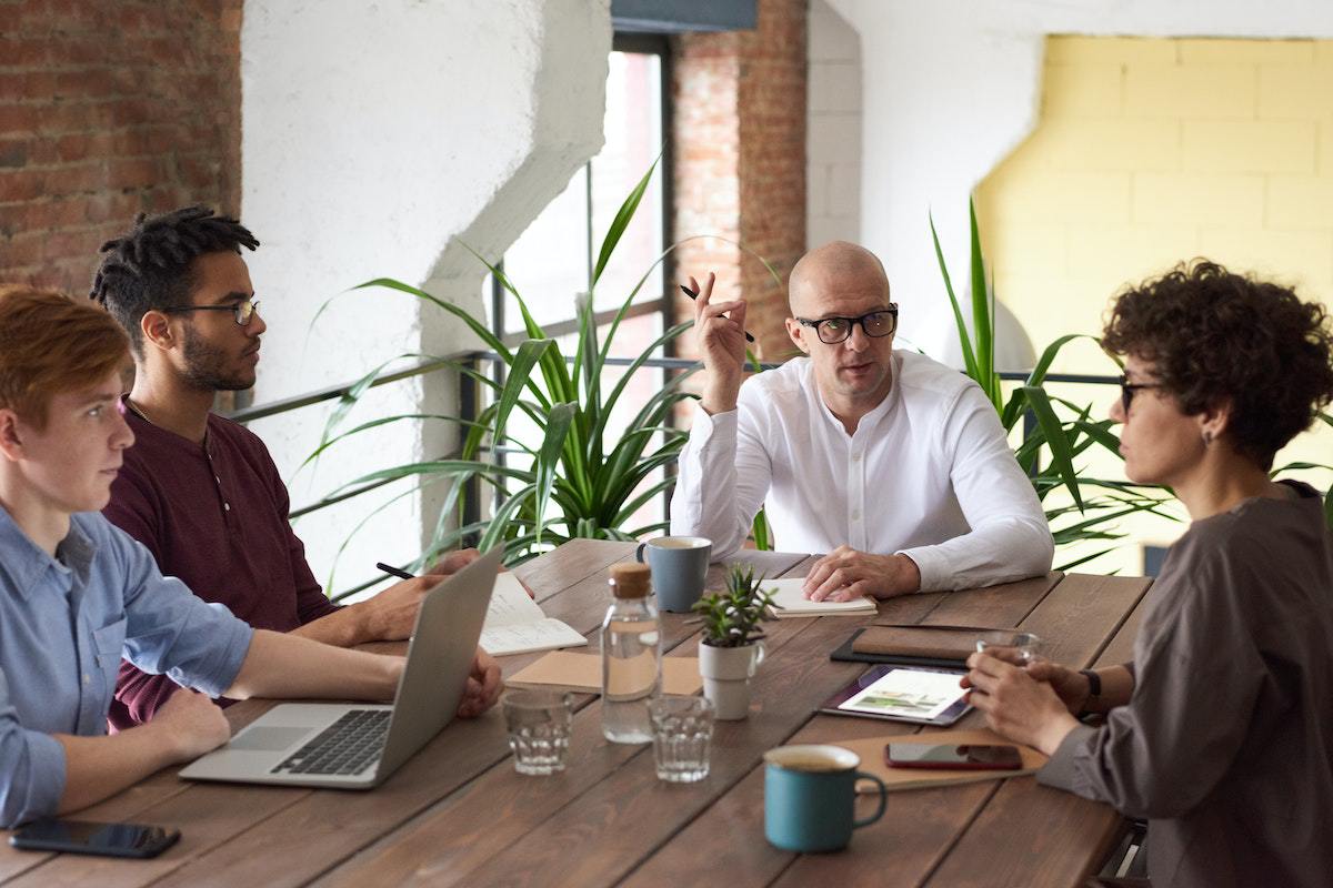 Três homens e uma mulher conversando diante de mesa de madeira em ambiente de reuniões de empresa debatendo tema e usando notebook e outros aparelhos
