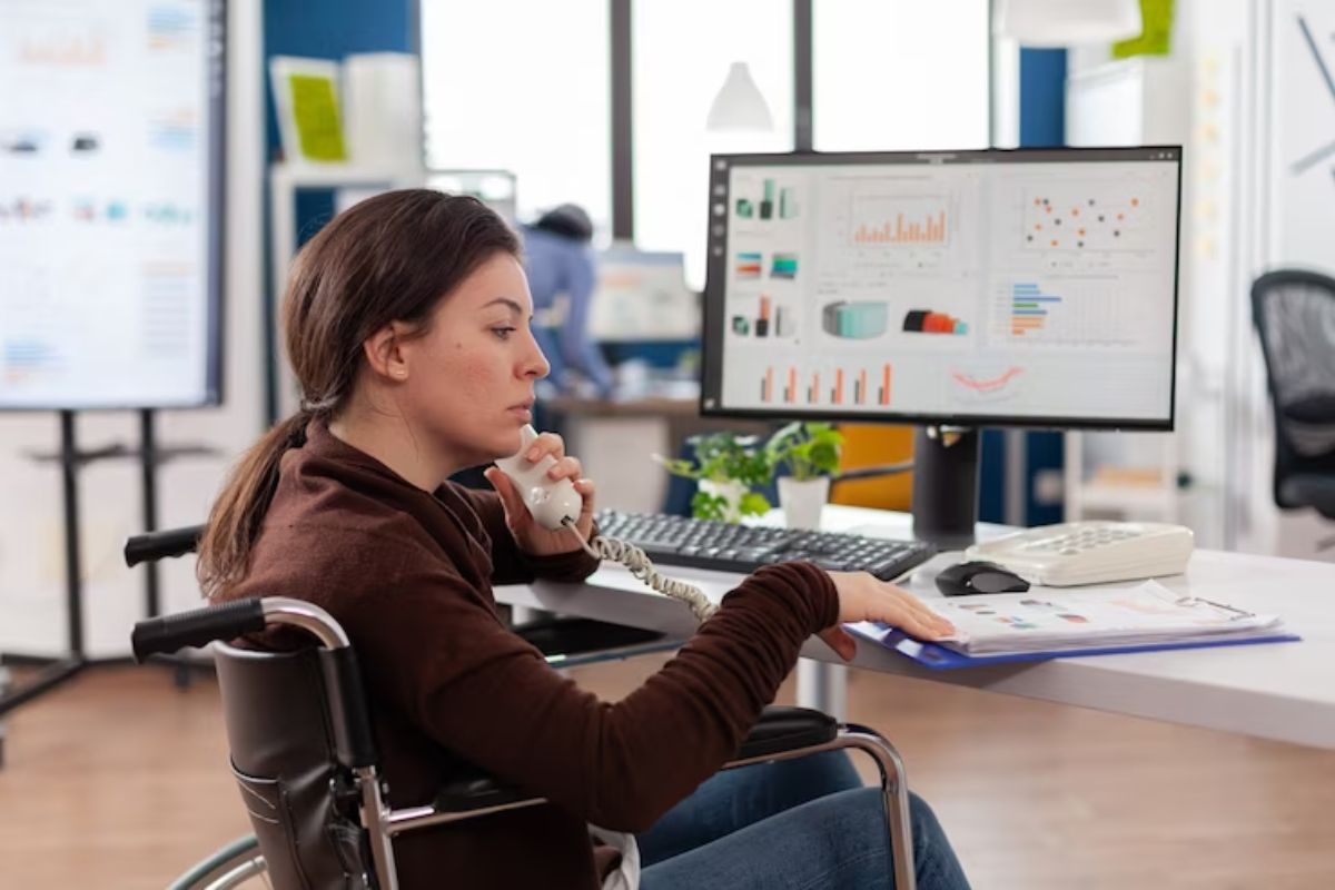 Mulher usando computador enquanto trabalha em escritório e fala ao telefone, em situação de cadeira de rodas, diante de mesa de madeira