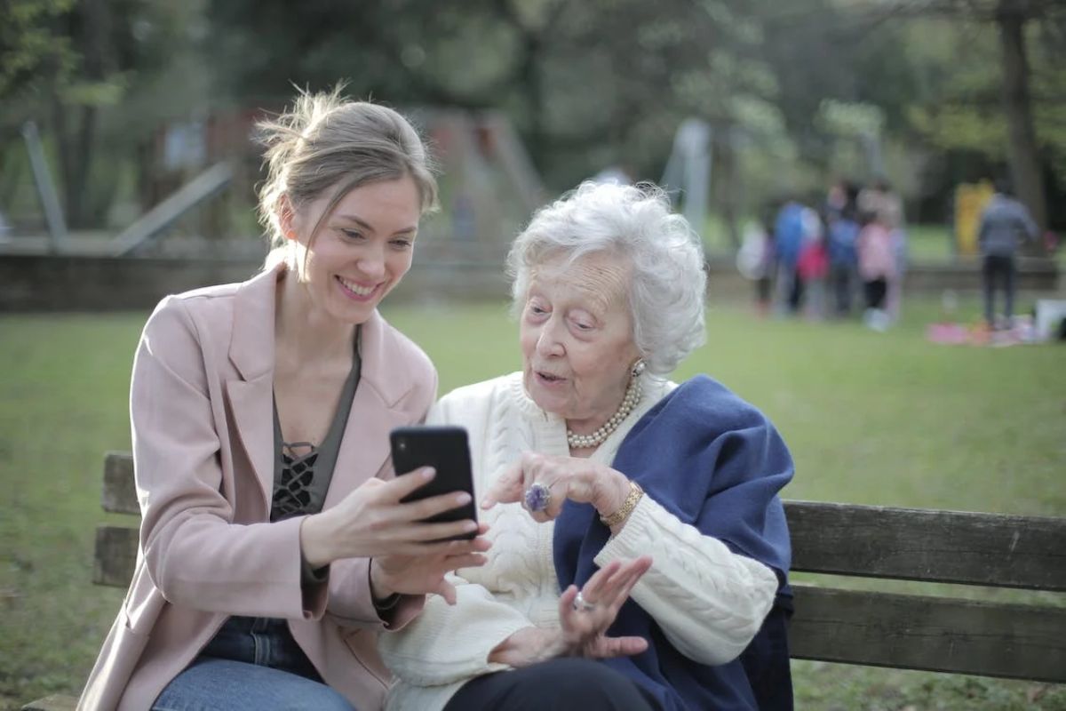 Mulher sentada ao lado de senhora em banco de parque mostrando algo à mesma em smartphone conectado à internet