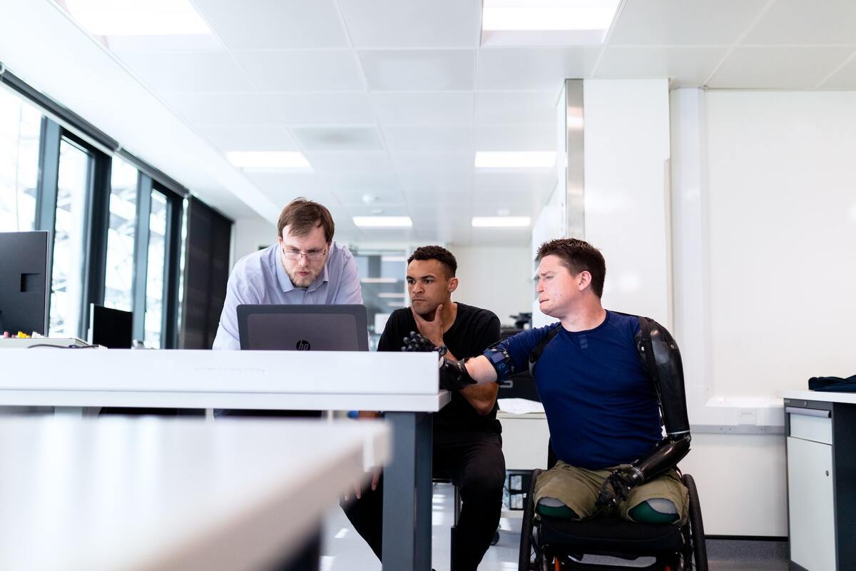 Homem em situação de cadeira de rodas em interior de ambiente de empresa com homem sentado à sua direita e outro de pé, enquanto analisam algo em notebook sobre mesa de trabalho alta