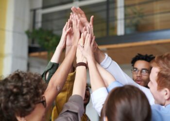 Pessoas em Ambiente de trabalho diverso tocando a palma das suas mãos sobre suas cabeças em sinal de comemoração e união da equipe