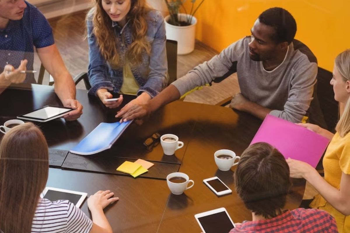 Equipe de trabalho diversa e inclusiva sentada ao redor de mesa redonda de madeira em empresa praticando a Comunicação em reunião