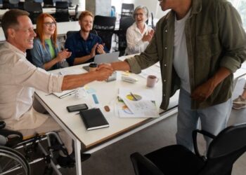 Homem de pé diante de equipe de trabalho diversa e inclusiva reunida ao redor de mesa em escritório enquanto cumprimenta homem em cadeira de rodas como demonstração de uma Comunicação saudável