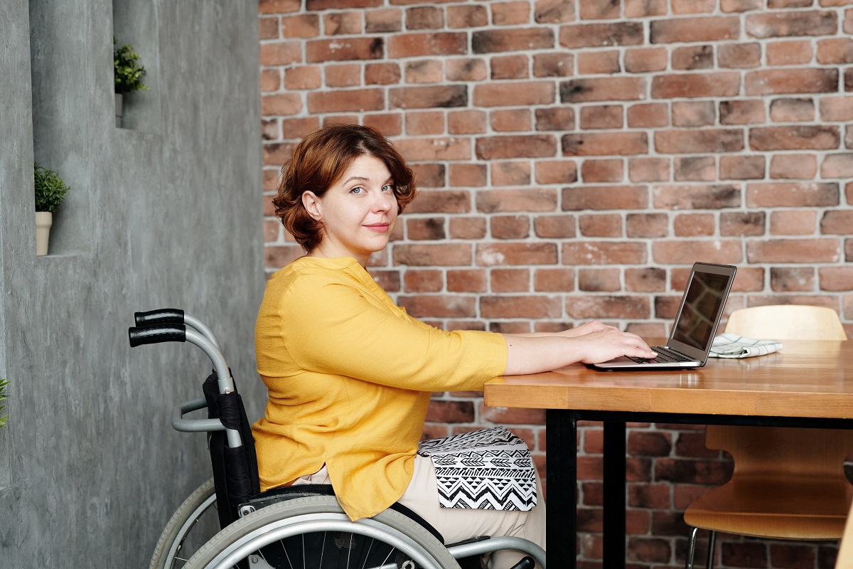 Mulher que usa cadeira de rodas usando notebook sobre mesa de madeira diante de parede rústica de tijolos sem reboco em empresa diversa que promove a inclusão por meio da Contratação de pessoa com deficiência
