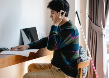 Homem sentado diante de madeira com folhas à sua frente, de olhos fechados e lendo escritos em Braille com sua mão direita, enquanto a esquerda vai ao ouvido, segurando fone tipo headset, que está conectado via fio a notebook também sobre a mesa, mas à direita do rapaz, com seu bastão do mesmo lado, porém apoiado na parede