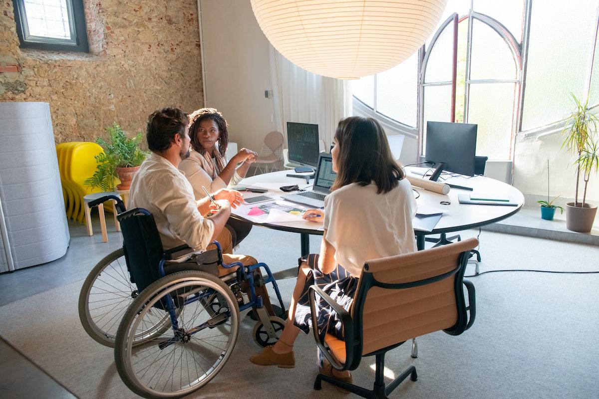 Homem em situação de cadeira de rodas em empresa com duas mulheres sentadas uma em cada lado ao redor de mesa redonda representando uma Equipe diversa e inclusiva