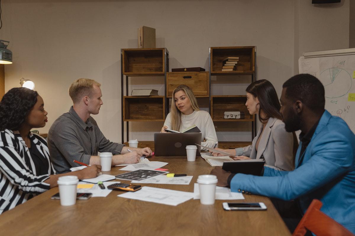 Reunião em local de trabalho ao redor de mesa de madeira em empresa com homens e mulheres de diversas raças e etnias debatendo Estratégia de diversidade e inclusão