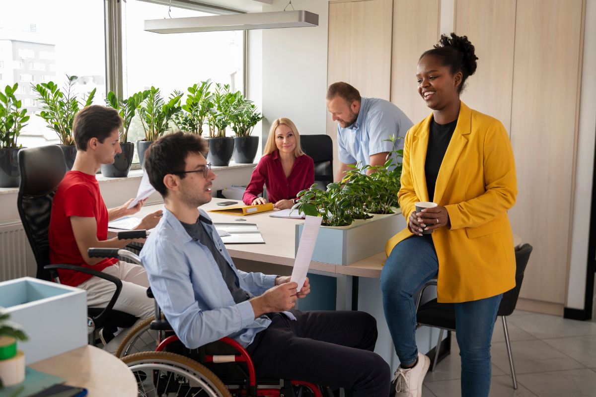 Homem em cadeira de rodas segurando papéis enquanto conversa com colegas homens e mulheres em escritório de empresa