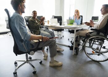 Local de trabalho diverso com homem em situação de cadeira de rodas conversando com outros dois homens e mulher sentados ao redor de mesa em empresa que considera que a Inclusão é o futuro