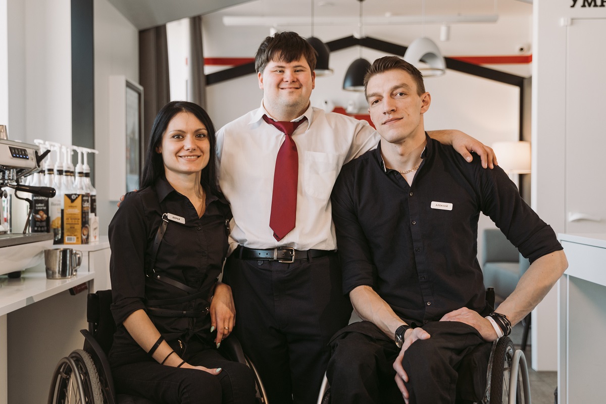 Plano de seleção inclusiva representado por homem de pé que apresenta síndrome de down abraçando homem e mulher em situação de cadeira de rodas em ambiente de empresa diversa