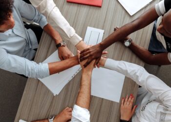 Treinamentos em equipe para melhoria de cultura com pessoas de etnias diferentes dando as mãos ao redor de mesa estreita de madeira com papéis sobre a mesma