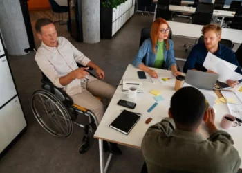 Um grupo de profissionais trabalhando juntos em uma reunião, incluindo um homem em cadeira de rodas, enquanto discutem estratégias empresariais com documentos e laptops em uma mesa.